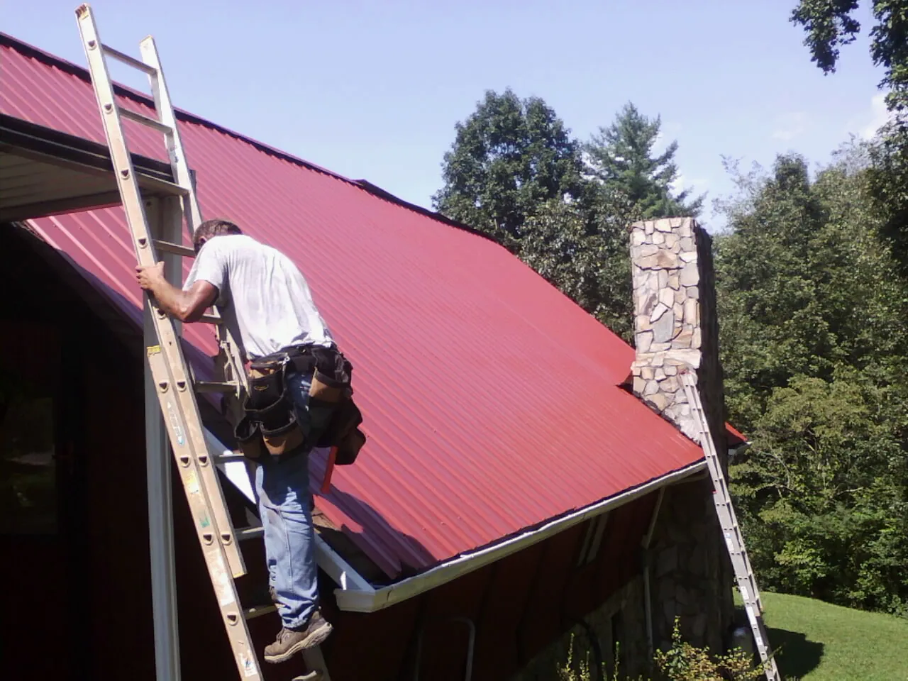 Roofer on a ladder beside a red metal roof and stone chimney — Soffit & Fascia Repair by Roofing Brothers in Adelphi